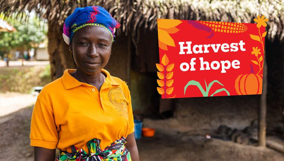 A woman in an orange shirt and patterned headwrap smiles slightly, standing outside her home, with a "Harvest of hope" sign to the right.