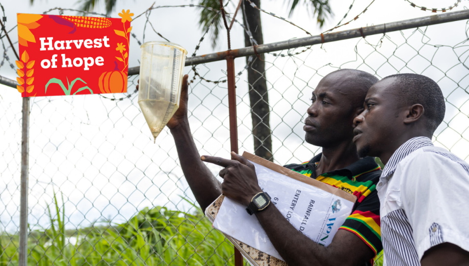 Two men examine a rainfall gauge next to a wire fence, with a colourful "Harvest of hope" sign beside them.