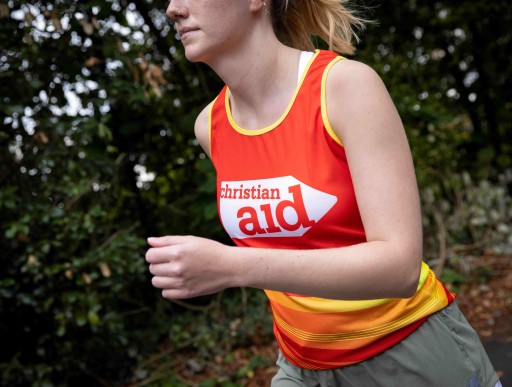 A woman running. There are trees behind her, and she's wearing and red and yellow Christian Aid running vest.