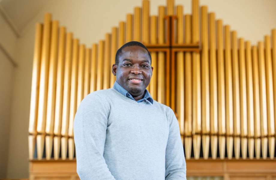 A man stands in a church smiling at the camera. Behind him is the pipes of an organ