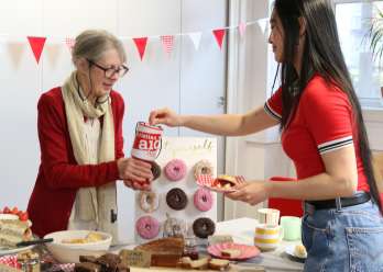 A woman holding a Christian Aid donation bucket accepting a donation from a supporter, stood behind a table full of baked goods. There is Christian Aid bunting behind her.